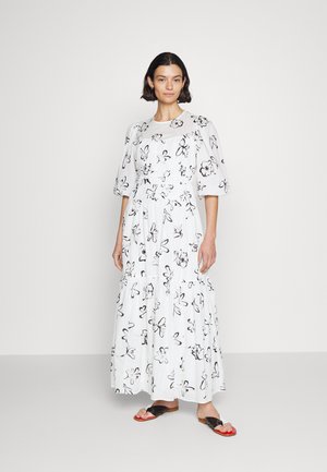 Robe blanche avec des imprimés floraux noirs, manches bouffantes, jupe à volants, col rond et coupe décontractée. Le mannequin porte des sandales plates noires et rouges.