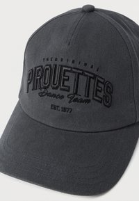 Harmaalippalakki, tehty tukevasta kankaasta, jossa kohokuvioitu "Pirouettes Dance Team" -teksti ja perustamisvuosi. Kaareva lippa.