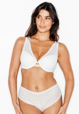 Donna che indossa un completo intimo in pizzo bianco con reggiseno e slip a vita alta, in piedi di fronte a uno sfondo bianco uniforme.