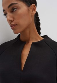 Black dress with a high neckline and a subtle V-cut. The fabric has a smooth texture, featuring structured shoulders and minimal detailing.