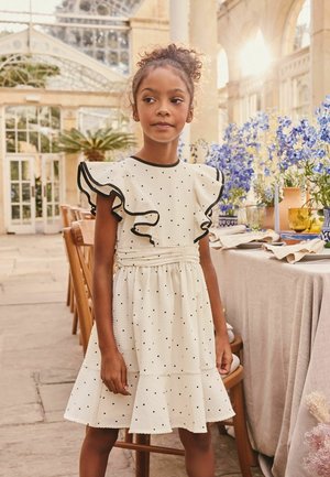 Fille aux cheveux bouclés portant une robe blanche à pois avec des manches à volants se tient à côté d'une table dressée avec des fleurs et de la verrerie dans un espace intérieur baigné de lumière.