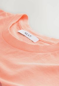  ferskfussisk T-shirt i ferskenfarvet bomuld med rund halsudskæring, ribbet krave og mærke med teksten "NEXT" i en hvid broderet etiket. Glat tekstur.