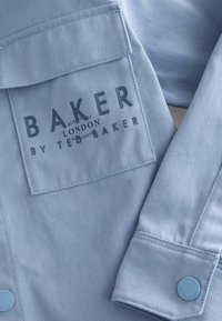 Svetlomodrá denimová košeľa s vreckom na hrudi. Obsahuje tmavý potlačený logo "BAKER BY TED BAKER" a dve modré gombíkové uzávery. Hladká textúra.