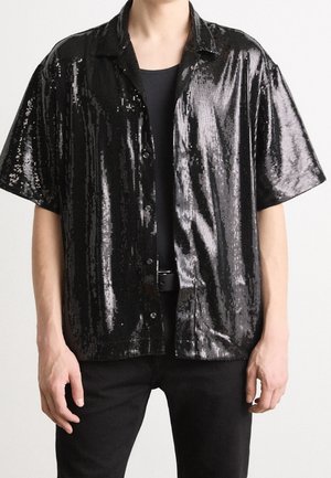 Mann trägt ein kurzärmliges schwarzes Paillettenshirt mit Knöpfen über einem schwarzen T-Shirt, mit schwarzer Hose und Gürtel vor einfachem Hintergrund.