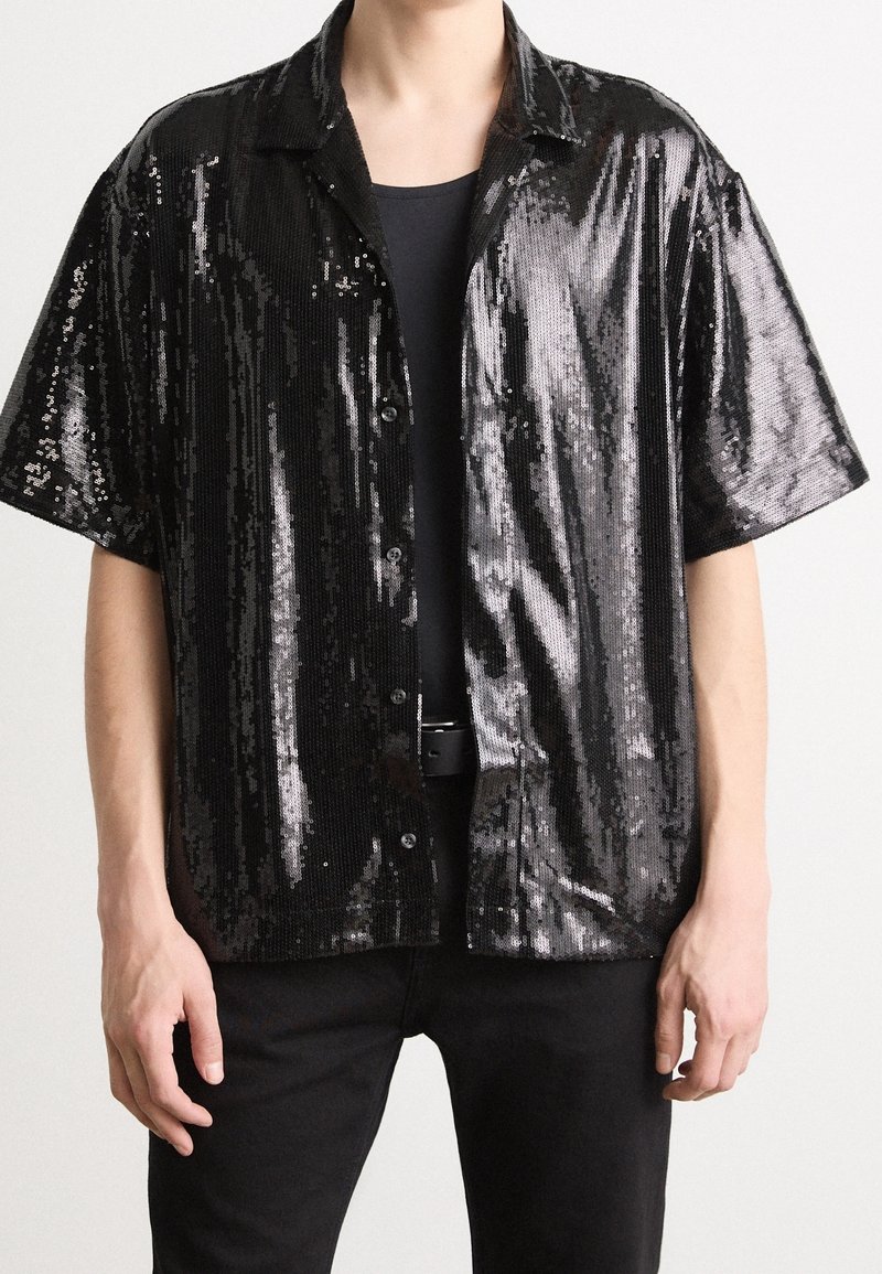 Homme portant une chemise noire à sequins à manches courtes boutonnée par-dessus un T-shirt noir, avec un pantalon et une ceinture noirs sur un fond uni.