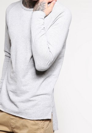 Chemise à manches longues gris clair en tissu doux, avec un col arrondi, des fentes sur les côtés et des manches ajustées. Surface texturée.