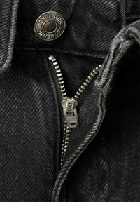 Material din denim negru, văzut în prim-plan, cu un fermoar metalic parțial deschis și un nasture inscripționat cu "Authentic Denim MNG".