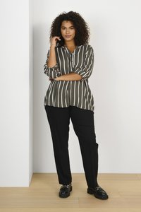 Blouse rayée vert olive avec des lignes verticales blanches et des manches retroussées, assortie à un pantalon droit noir et des chaussures noires à semelles épaisses.