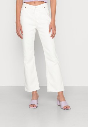 Jeans brancos de perna larga com um acabamento texturizado, apresentando um fecho de botão e bolsos, combinados com sandálias lilás de salto bloco que se calçam facilmente.