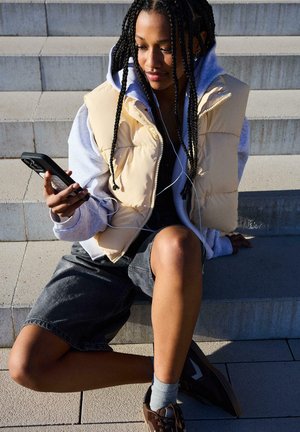 Mlada ženska s pletenicami, oblečena v bež telovnik in sivo kapucarico, sedi na zunanjih stopnicah in gleda v svoj pametni telefon z vstavljeno slušalko.