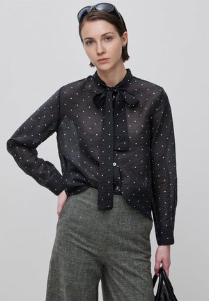 Jeune femme aux cheveux courts portant un chemisier noir transparent à pois, un pantalon gris à carreaux, des lunettes de soleil sur la tête, tenant un sac à main noir.