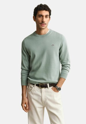 Mann mit kurzem lockigem Haar und Schnurrbart, trägt hellgrünen Pullover, weiße Hose, braunen geflochtenen Gürtel und Armbanduhr, steht mit der Hand in der Tasche.