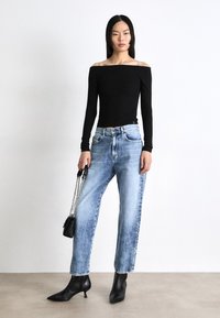 Juodas off-the-shoulder ilgarankovis megztinis derinamas su šviesiai mėlynomis laisvo kirpimo džinsomis. Modelis vilki juodas aulinukus su smailiu priekiu ir laiko mažą juodą rankinę.