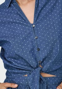 Blå denimskjorta med krage, knapplist fram, knytmidja och subtila geometriska mönster genom hela tyget.