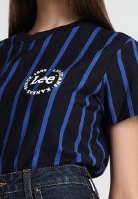 Czarna koszulka z niebieskimi pionowymi paskami, z białym okrągłym logo na środku. Krótkie rękawy i okrągły dekolt.