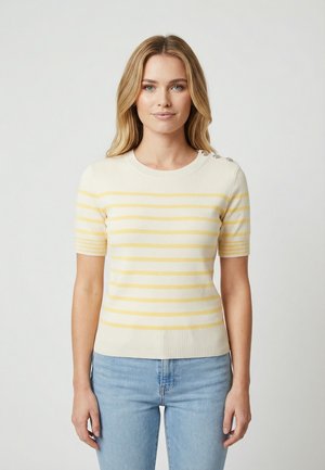 Femme aux cheveux longs et blonds portant un pull à manches courtes rayé crème et jaune pâle avec des boutons sur l'épaule et un jean bleu clair.