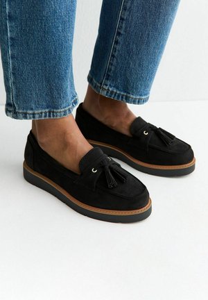 Pieds portant des mocassins à pampilles en daim noir avec des semelles marron, associés à un jean bleu raccourci sur un fond blanc.