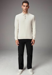 Camisa polo de manga larga color beige claro con cuello y tres botones, combinada con pantalones negros y zapatillas blancas, de pie sobre un fondo neutro.