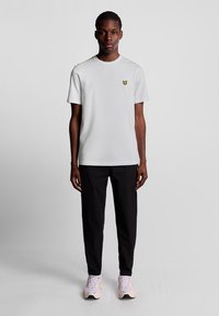 Ung mand står, iført en hvid t-shirt med gul logo, sorte bukser og lyserøde sneakers, foran en ensfarvet grå baggrund.