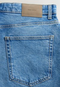 Jasnoniebieskie dżinsy z denimu mają prostą tylną kieszeń oraz prostokątną skórzaną metkę z wytłoczonym napisem "DENIM SUPPLY" w ciemnych literach.