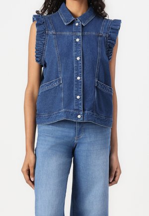 Veste sans manches en denim foncé avec boutons, emmanchures volantées et poches avant, portée avec un jean en denim bleu clair.