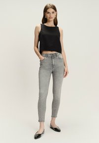 Kobieta stoi w bezrękawnikowej czarnej bluzce crop top, szarych obcisłych spodniach z wysokim stanem, czarnych balerinach i srebrnych kolczykach w kształcie kółek na tle jednolitym.