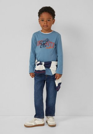 Blaues Langarmhemd mit einem Dinosaurier-Design, blaue Jeans und weiße Sneakers mit beigen Akzenten; gestreifter Pullover um die Taille gebunden.