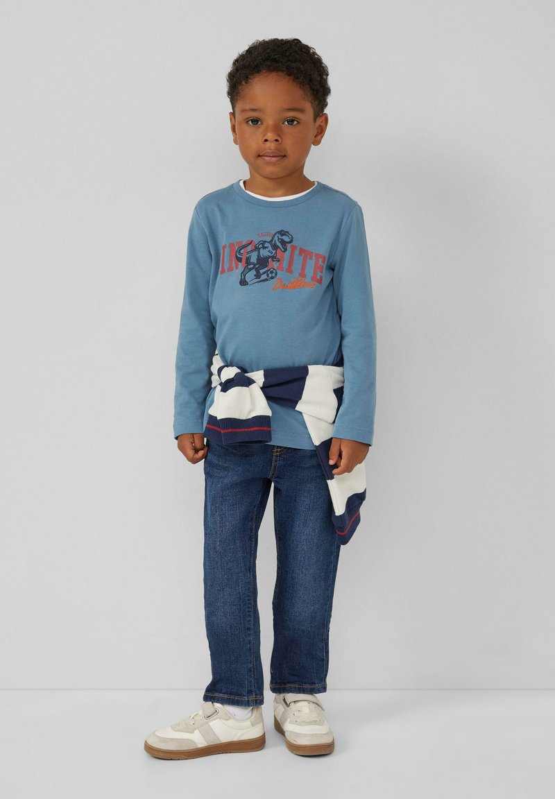 Blaues Langarmhemd mit einem Dinosaurier-Design, blaue Jeans und weiße Sneakers mit beigen Akzenten; gestreifter Pullover um die Taille gebunden.