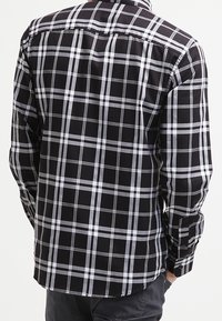 En person står vänd bort, iklädd en svart och vit rutigt skjorta, vilket visar plaggets baksidedesign och åtsittande siluett.