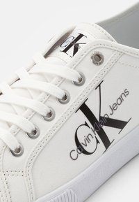 Biały tenisówka na płótnie z czarnym logo. Posiada zapięcie na sznurowadła, srebrne przelotki i teksturowaną białą gumową podeszwę. Prosty design i wyraźne logo.