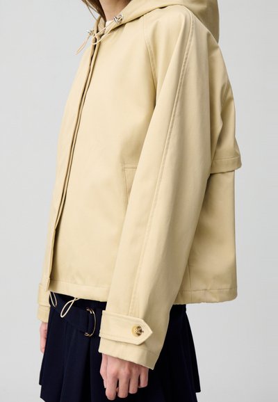 Veste à capuche beige fabriquée en matériau léger et lisse. Elle dispose d'un cordon de serrage, de poches latérales et de poignets à boutons.