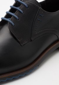 Chaussure en cuir noir avec tige en maille texturée, lacets bleus, semelle extérieure marron contrastée et motif en zigzag sur le bord de la semelle.