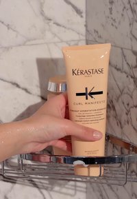 Kérastase Curl Manifesto conditioner in een perzikkleurige tube, vastgehouden in een natte hand, met zwarte tekst en metallic accenten op een chroomrek.
