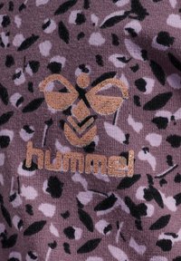 Lila Stoff mit einem schwarz-weißen Blattmuster, ausgestattet mit einem glänzenden goldenen Bienenlogo und dem "hummel"-Text in passender goldener Farbe.