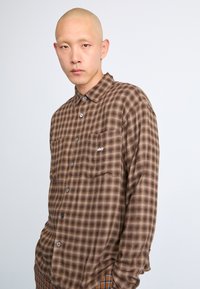Brown plaid button-up shirt with two front pockets, featuring a small embroidered logo. Soft fabric and relaxed fit.