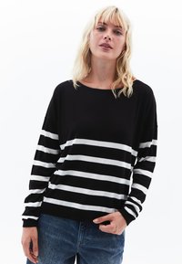 Schwarz-weißer gestreifter Pullover mit langen Ärmeln, rundem Ausschnitt und geripptem Saum; kombiniert mit hellblauen Jeans.