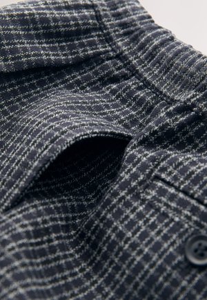 Marinblå texturerat tyg med rutnätsmönster, utrustat med elastisk midjeresår och knappdetalj. Närbild på invecklad väv och design.