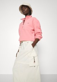 Sudadera de forro polar en rosa claro con media cremallera, con un pequeño logo, combinada con una falda larga color crema con bolsillos tipo cargador y cordones.