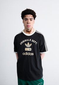 Melna kokvilnas T-kreklu ar zelta krāsā izšūtiem uzrakstiem "ORIGINALS DEPT." un "adidas". Kontrasta balti apkakles un piedurkņu akcenti, īpašas svītrotas plecu daļas.
