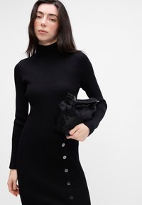 Čierne rebrované šaty s vysokým golierom, s bočnými gombíkmi. Model drží malú čiernu kožušinovú kabelku s striebornými kovovými detailmi.