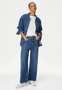 Chaqueta de mezclilla, camisa blanca con puntos amarillos, vaqueros de pierna ancha azules y zapatillas blancas. El conjunto presenta un ajuste relajado y un look en capas.
