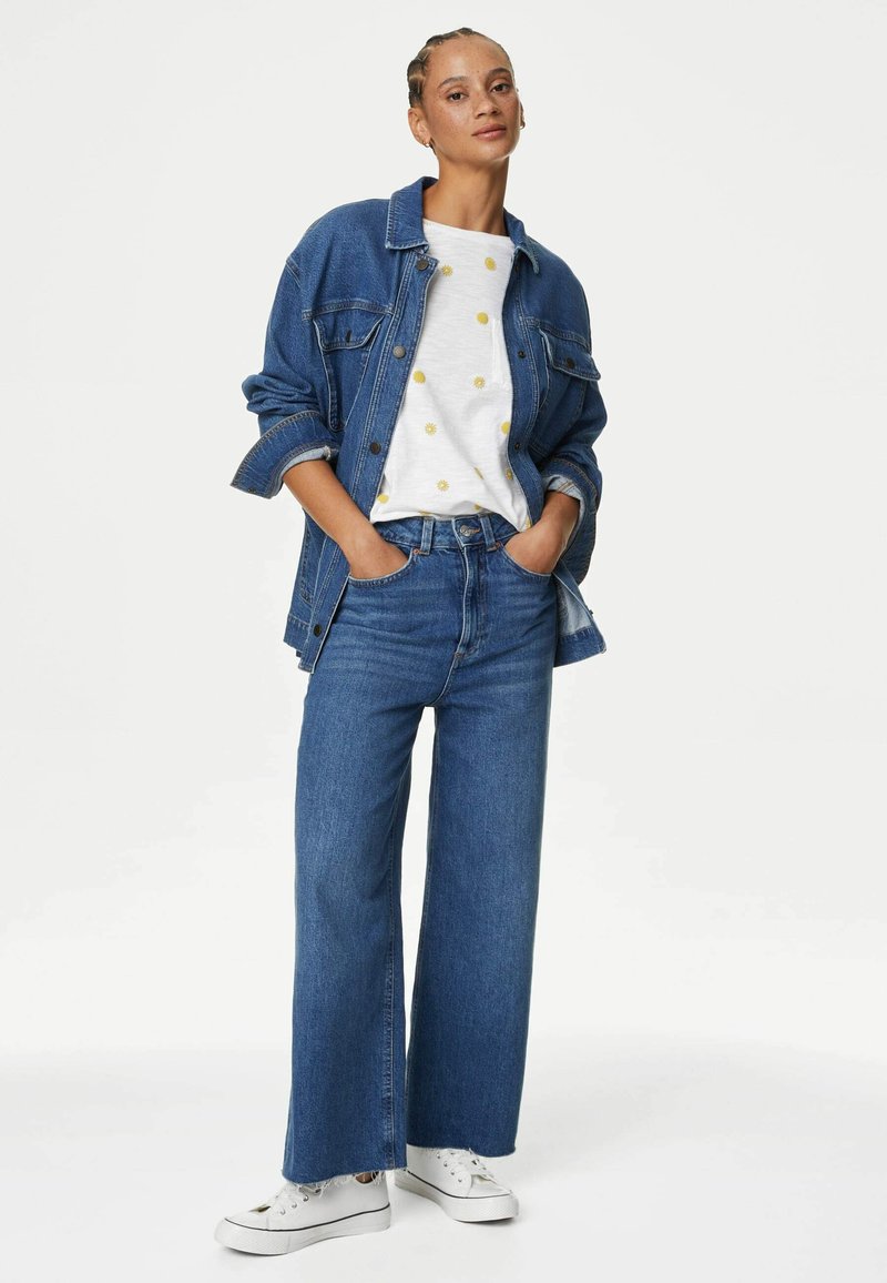 Chaqueta de mezclilla, camisa blanca con puntos amarillos, vaqueros de pierna ancha azules y zapatillas blancas. El conjunto presenta un ajuste relajado y un look en capas.