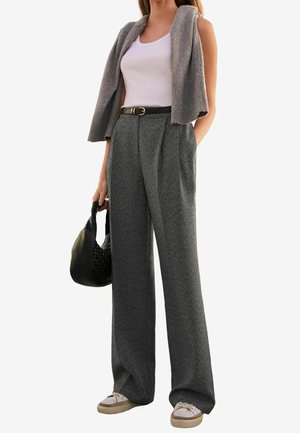 Kvinde iført grå vidde bukser, hvid tanktop, grå sweater lagt over skuldrene, hvide sneakers, holder sort flettet håndtaske.