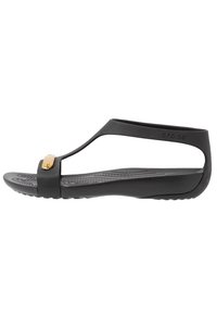 Musta kumisaapas, jossa on T-nauha design, kultainen korostus yläosassa ja karhennettu pohja pitävyyden vuoksi. Litteä pohja, minimalistinen tyyli.