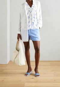 Chemisier blanc transparent boutonné sur un chemisier bleu à fleurs, associé à un short bleu clair et des sandales à talons transparentes. Tient un sac à main crème.