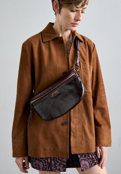 Veste en daim marron sur une robe à motifs, avec un sac en cuir marron foncé portant une fermeture éclair dorée et un logo, porté en bandoulière.