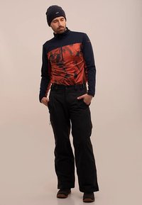 Langærmet overdel med lynlås i mørkeblå og orange med abstrakt mønster; sorte, tætsiddende bukser; sort hue; brune sko.