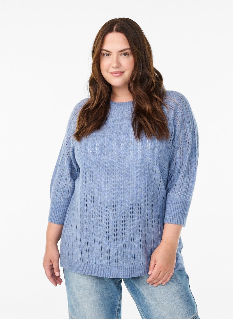 Frau mit langen braunen Haaren, die einen hellblauen, strukturierten Strickpullover und hellblaue Jeans trägt, steht vor einem weißen Hintergrund.