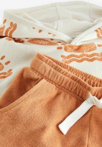 Hættetrøje i creme med orange abstrakte mønstre, parret med terracotta-farvede joggers med elastisk talje og snor.