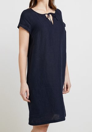 Vrouw draagt een loszittende donkerblauwe jurk met korte mouwen, een kleine V-hals en strikdetail bij de halslijn.